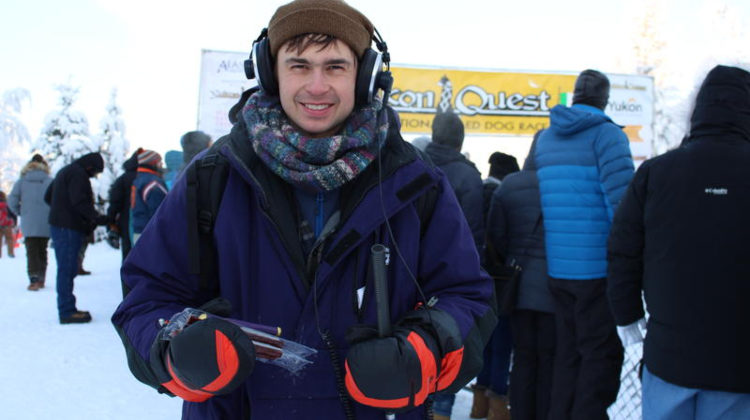 Man in winter gear with headphones and microphone near the start line of a dog race. Other people in winter gear are behind him.