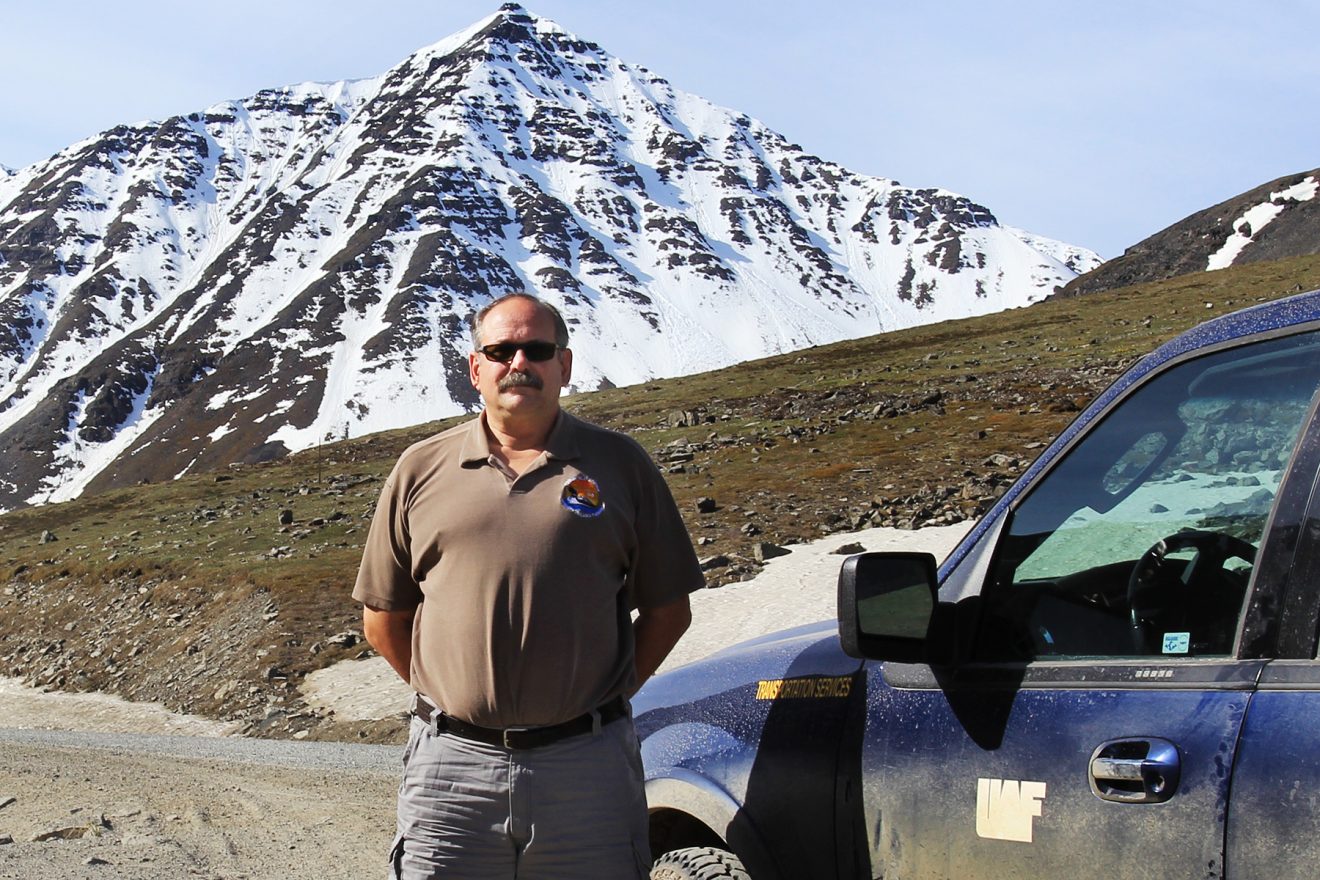 Man standing on a gravel road by a dusty truck in front of a snow-covered mountain.