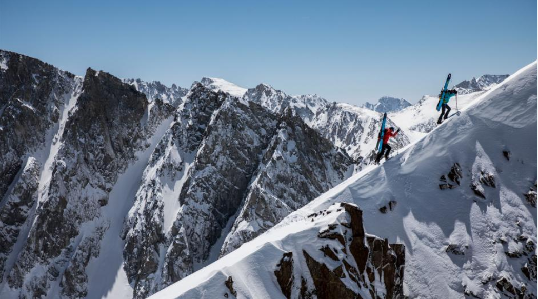 Photo of two snowboarders climbing a steep mountainside, surrounded by other mountains.