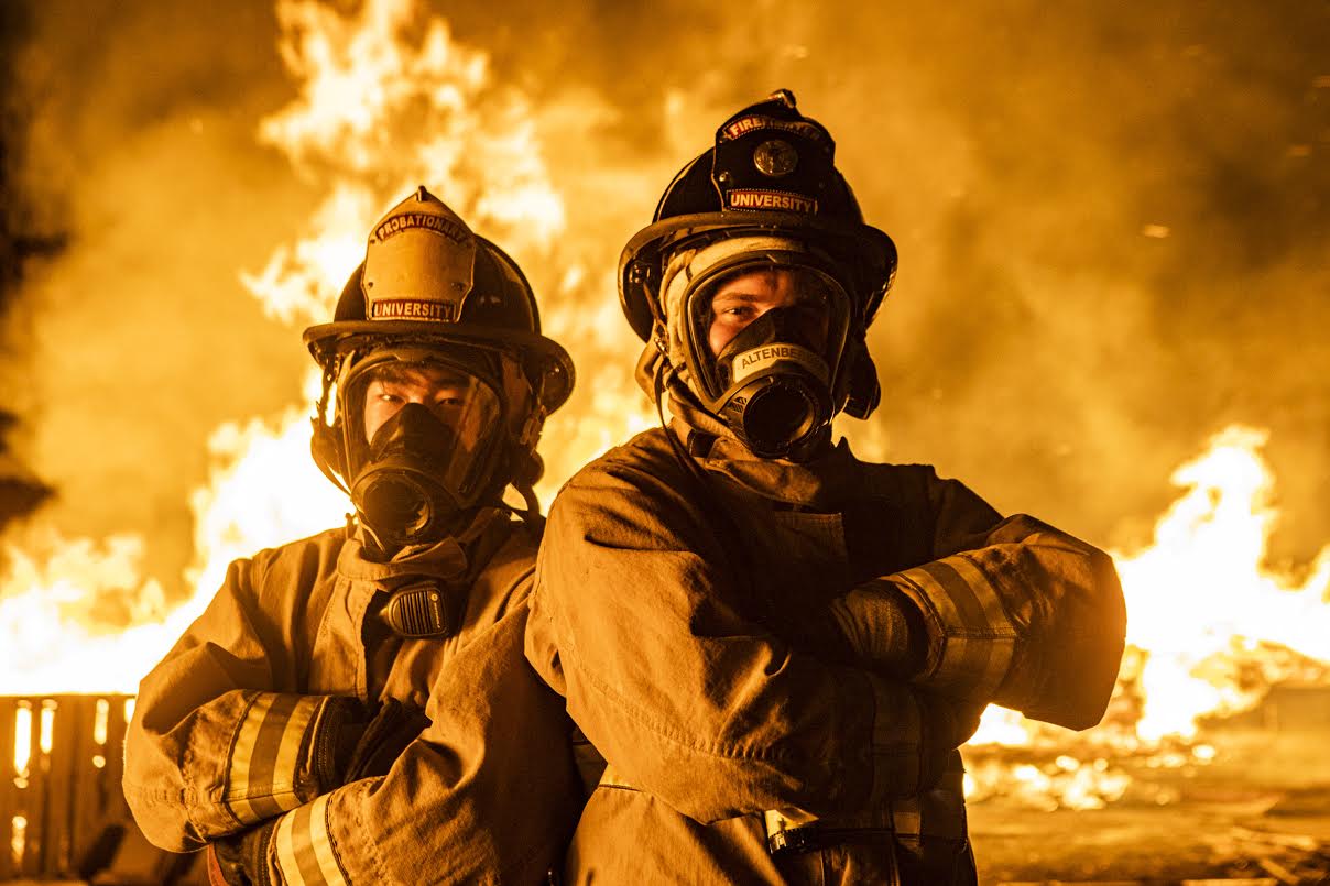 Two student firefighters in gear and masks stand, arms folded, in front of the bonfires during Starvation Gulch 2019.