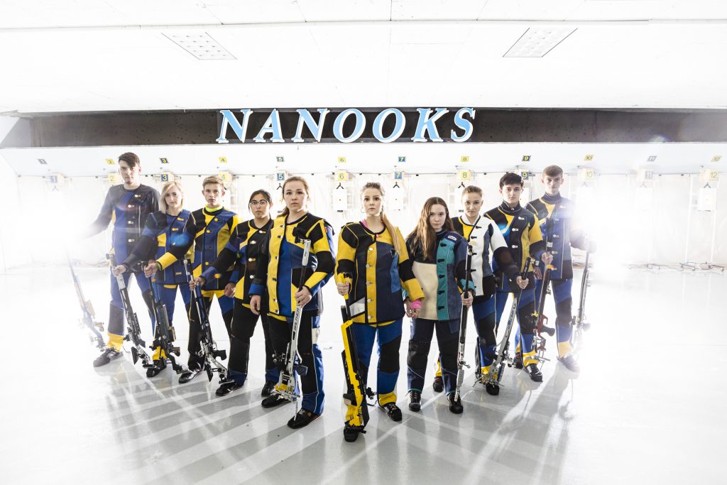 Members of the Nanooks rifle team stand in their gear and with their rifles in a v shape in the shooting range at UAF.