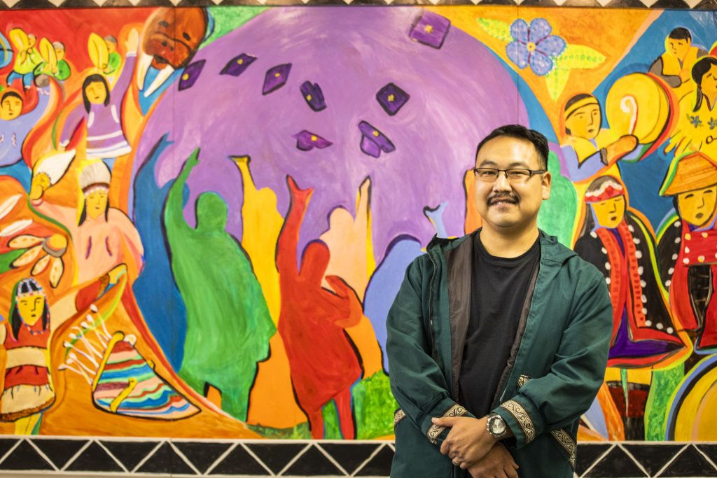 Artist Percy Avugiak stands in front of a brightly colored mural depicting figures from many Alaska Native traditions.