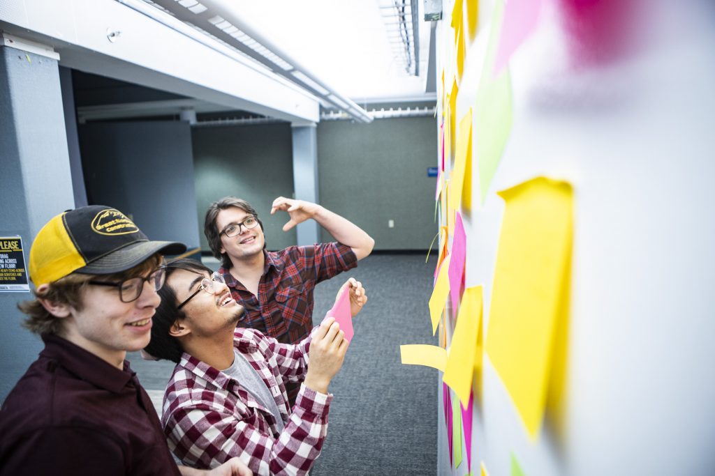 Three people stand at a whiteboard covered in pink and yellow stickies.