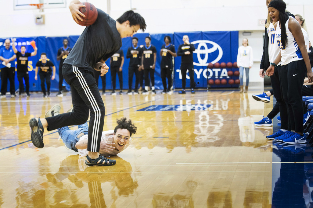 One student holds the ball high while another falls on the floor. Both are laughing. Members of the women's and men's basketball team stand on the sidelines cheering them on.