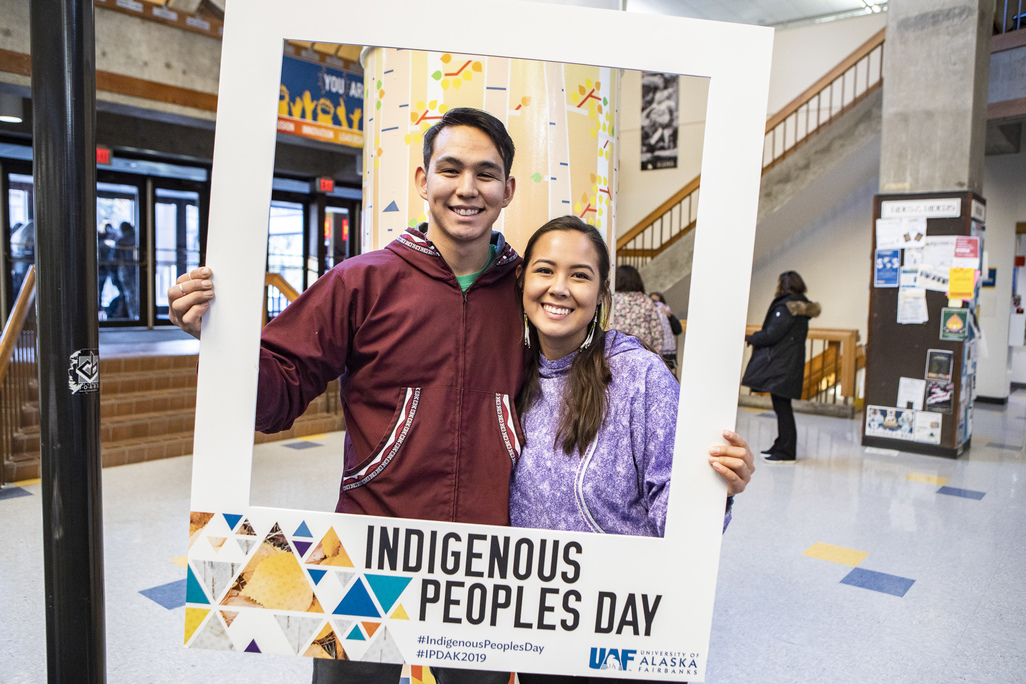Two students stand side by side holding a cardboard frame with Indigenous People Day graphic image painted on the bottom f it. They are in Wood Center.