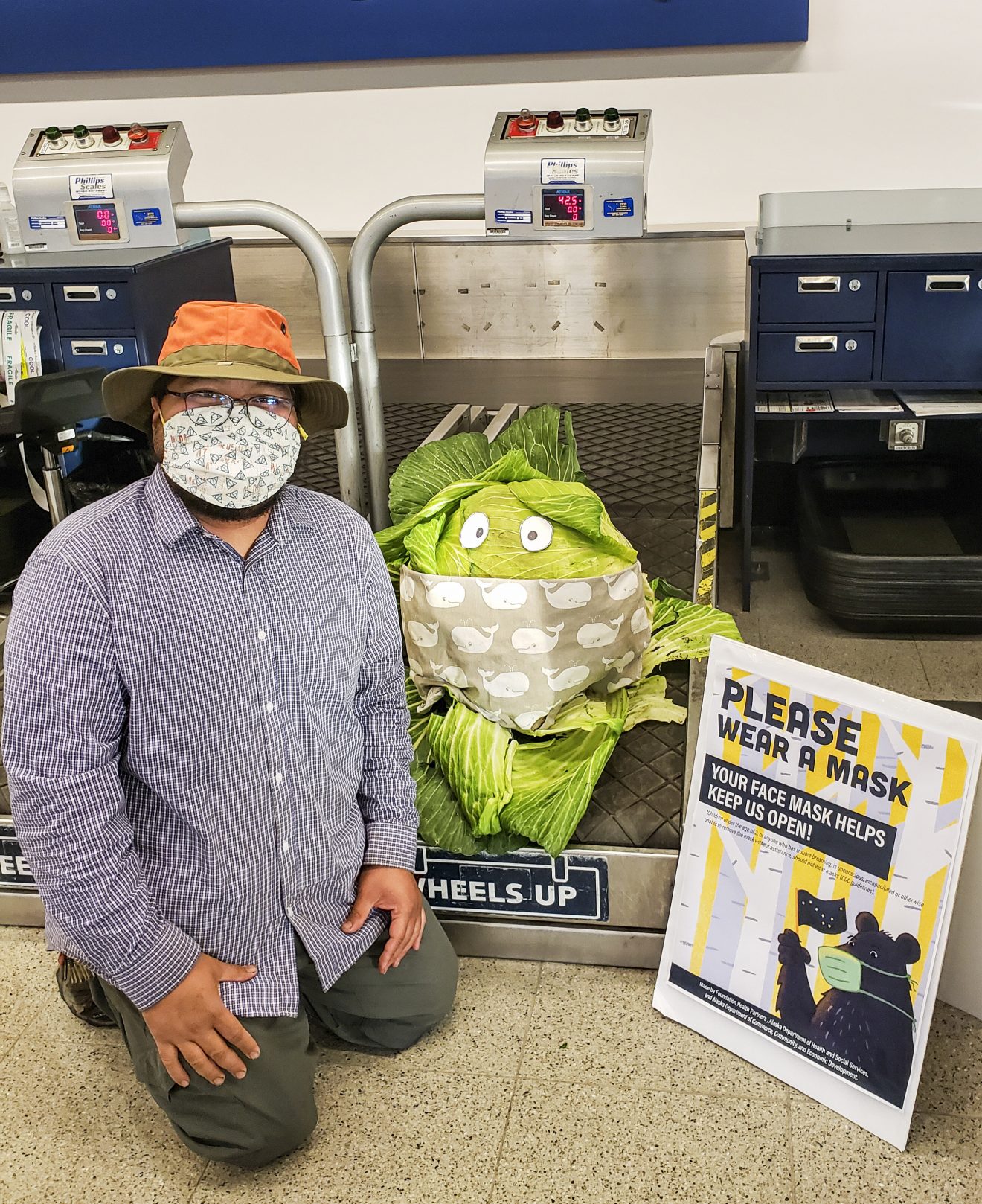 A large cabbage with googly eyes and a mask sits on an airport luggage scale, next to the man who grew it, also wearing a mask.