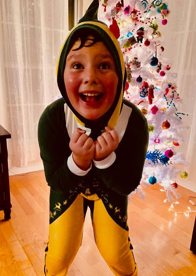 An excited young child in an elf costume standing in front of a Christmas tree.
