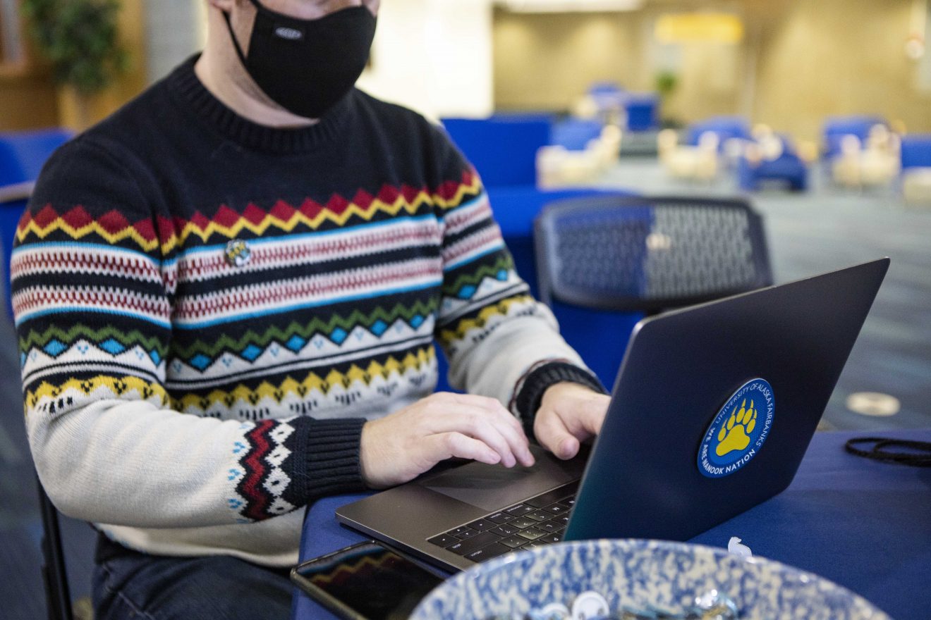 A man types on his laptop. He is wearing a mask.