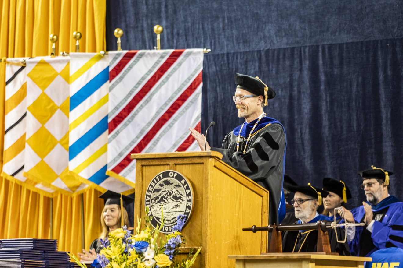 Man stands at podium, dressed in cap and gown. Flags hang behind him.