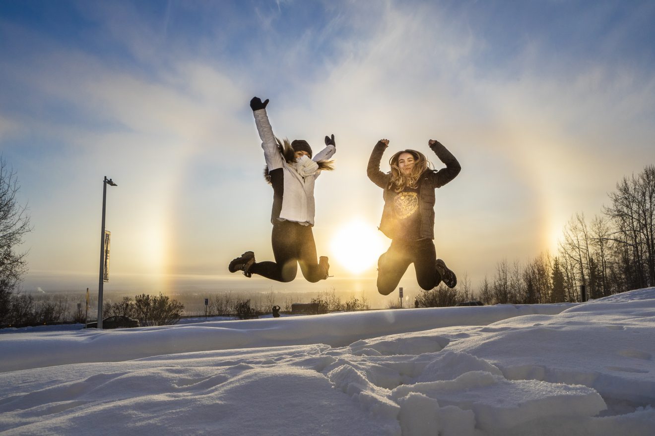 Two young women jump in the air. Snow covers the ground. The sun is low on the horizon even at midday.