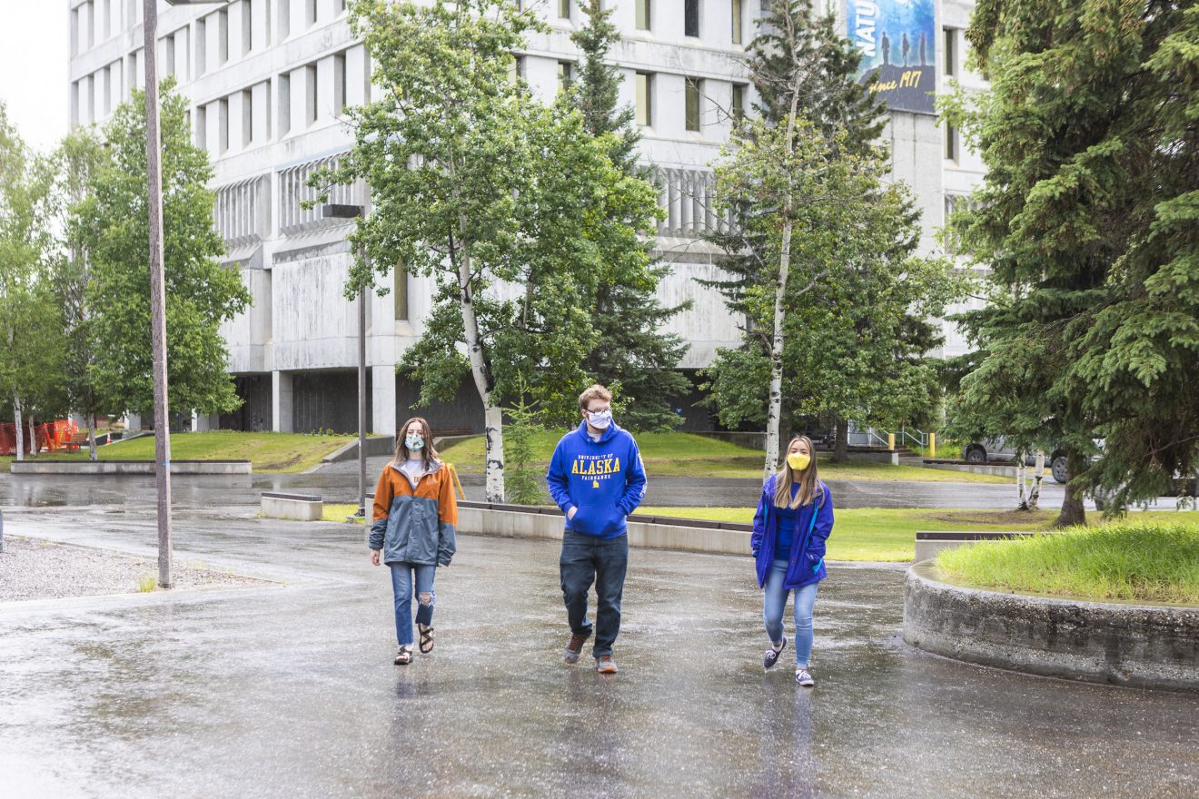 Three students wearing masks walk near a building. It is summer. The pavement is wet from a recent rain.