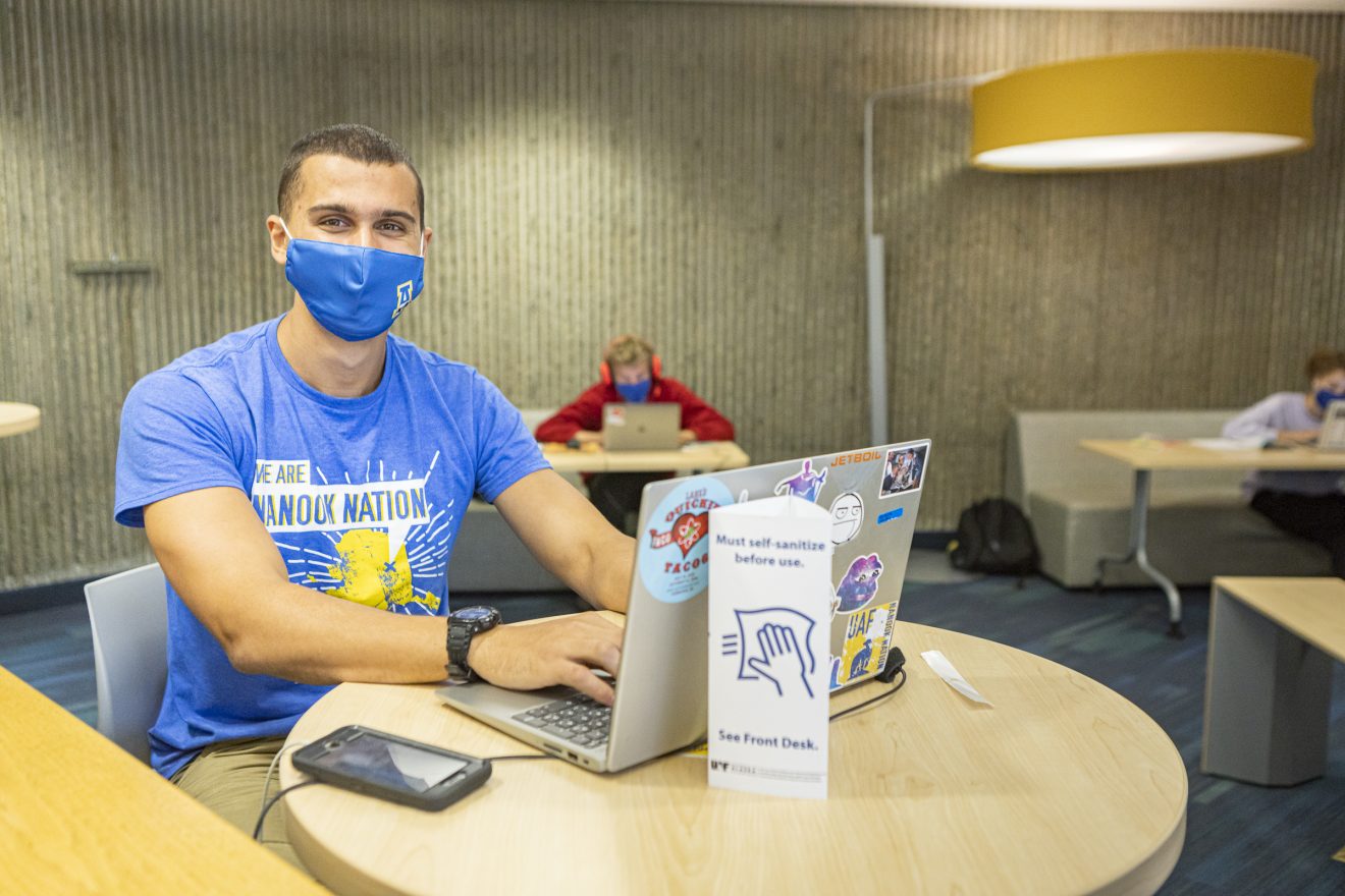 Student wearing UAF COVID mask sits at a round table in front of a laptop. Other students sit behind him, also wearing masks and working on laptops.