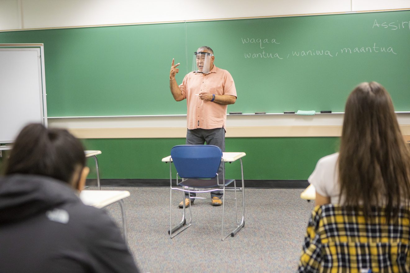 A professor wearing a face shield stands in front of a blackboard with some Yup'ik phrases written on it. Two students are within view.