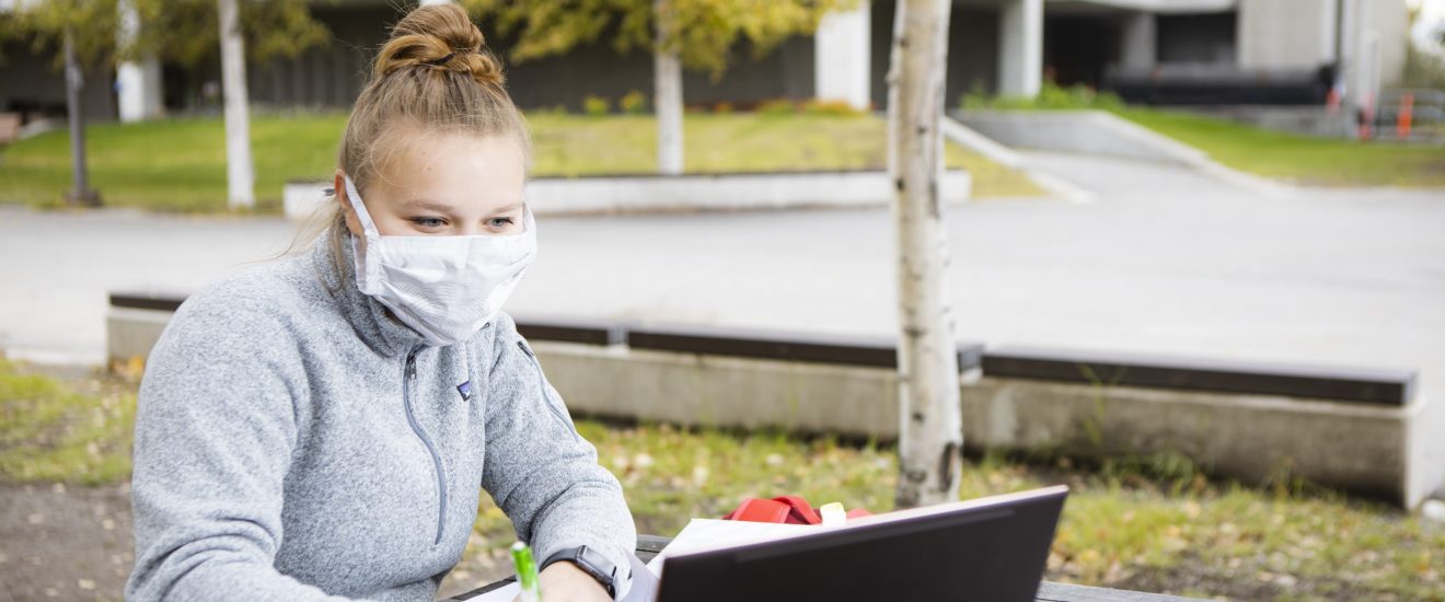 A student wearing a mask and a fleece sweater studies at her laptop outside. It is early fall.