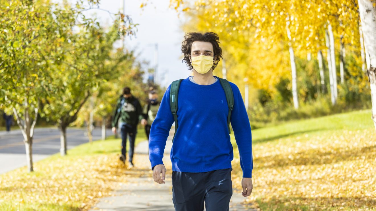 A man in a blue sweater and yellow mask walks along a sidewalk. It is fall.