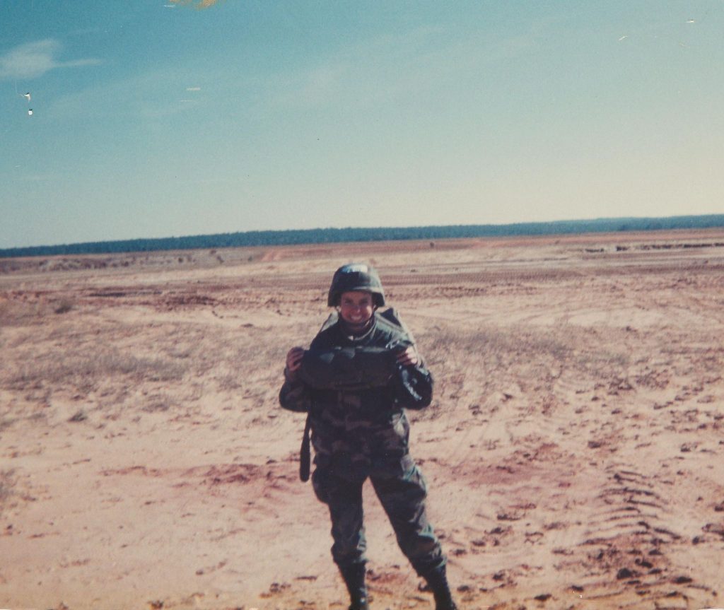 Woman in an Army jump suit and helmet standing on a large sandy field with a row of trees in the distance.