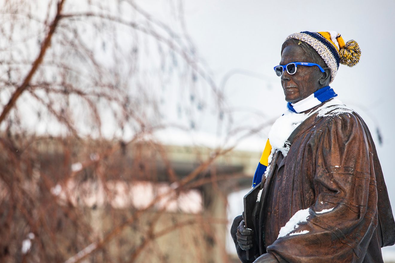 Statue of Charles Bunnell wearing a hat, scarf and sunglasses, all in blue and gold. He is partly covered in snow.