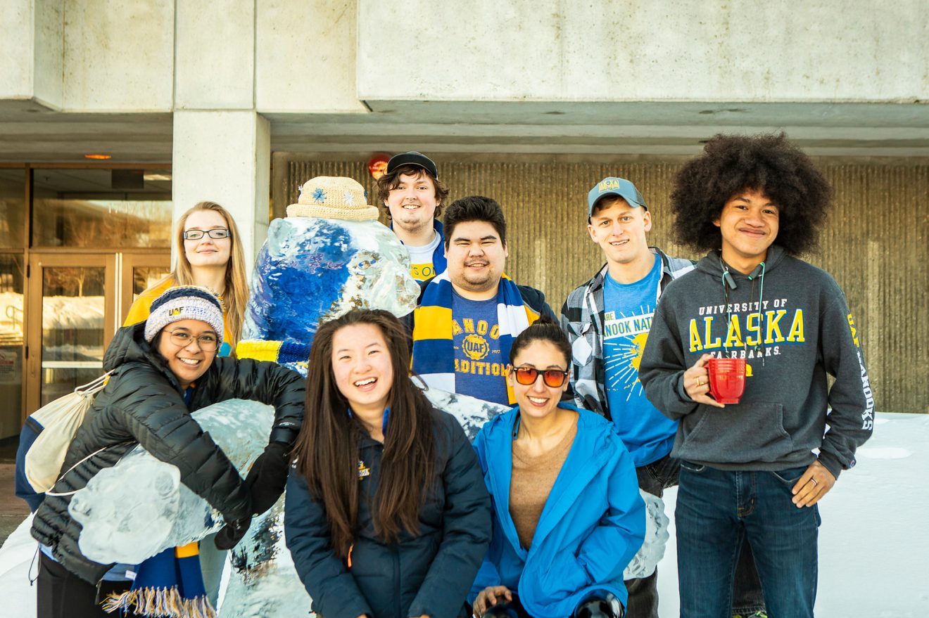Eight students stand around an ice sculpture of a polar bear outside a building. They are dressed in blue and gold or other UAF clothing.