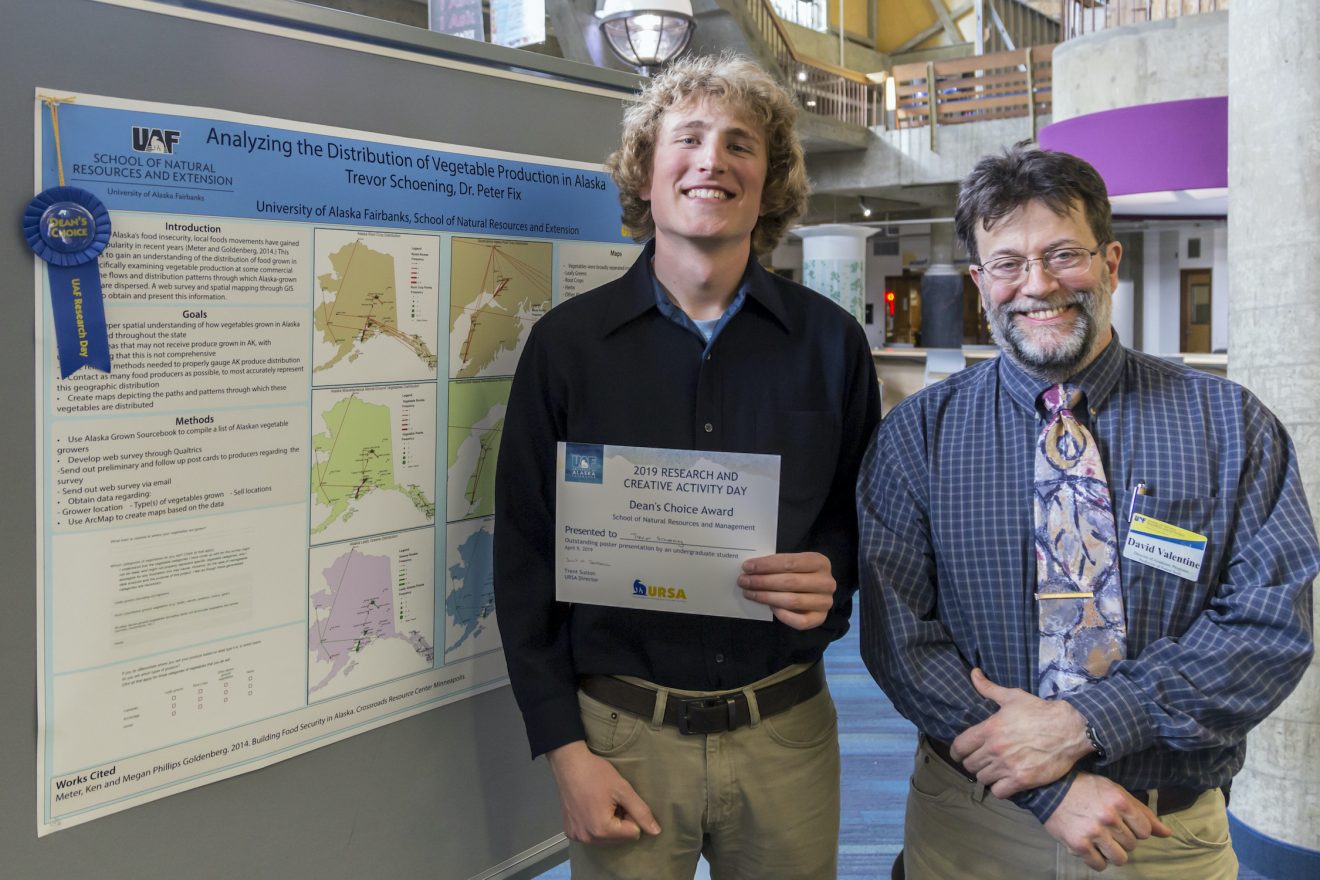 Two people stand beside a poster hanging on a board. The student is holding a certificate. The poster has a blue ribbon attached to it.
