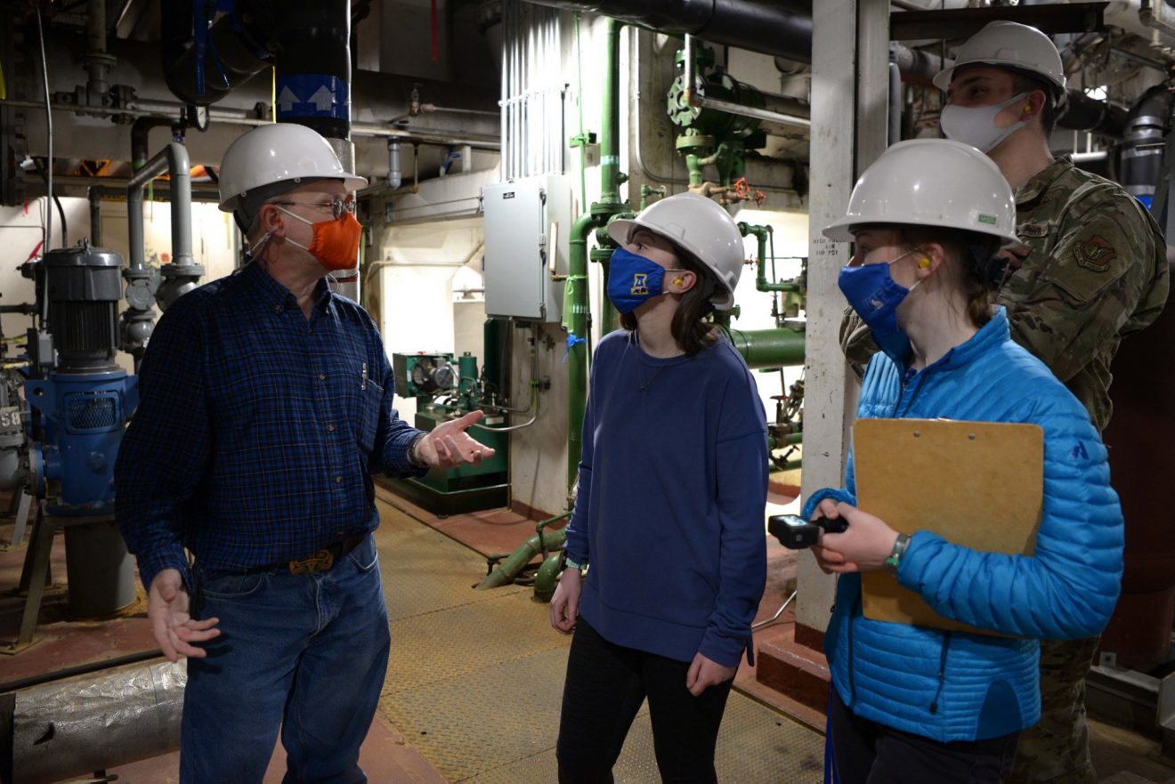 Two men and two students wearing hard hats stand in a large mechanical room. They are all wearing masks.
