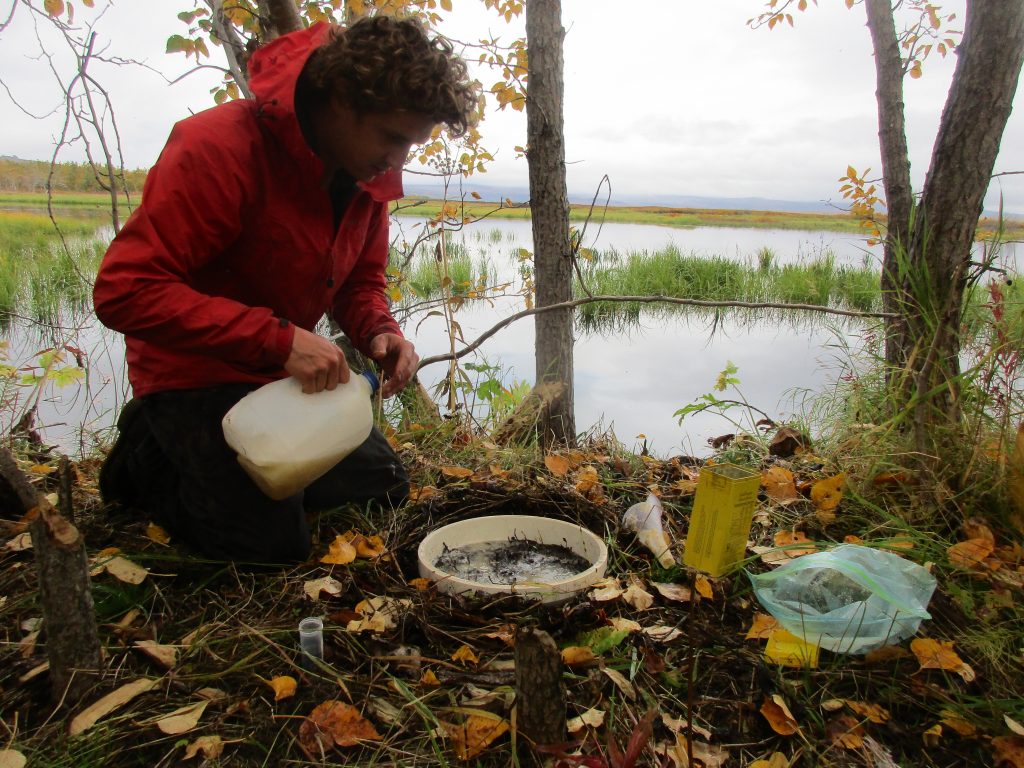 A man in a coat kneels on the ground. He's holding a milk jug. Various containers are on the ground in front of him. He's by a lake, and it is fall.