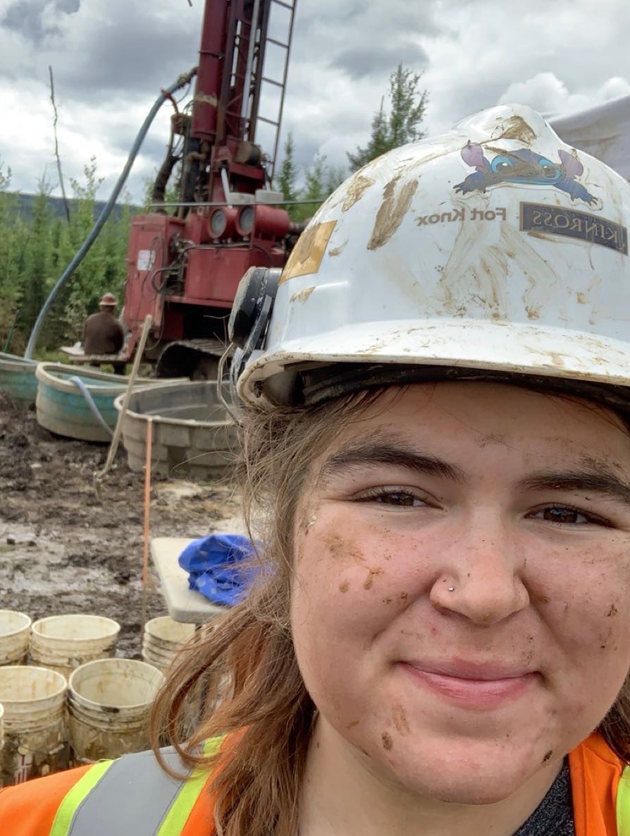 Closeup of a young woman wearing a hard hat and standing in front of equipment and buckets in a muddy area.