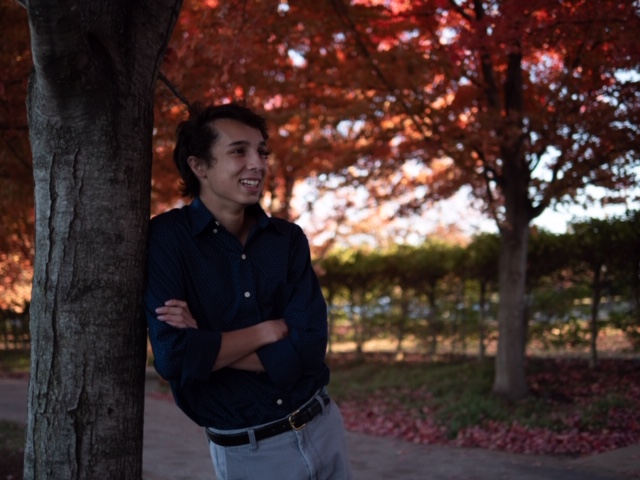 Photo of young man leaning against a tree. It is fall.