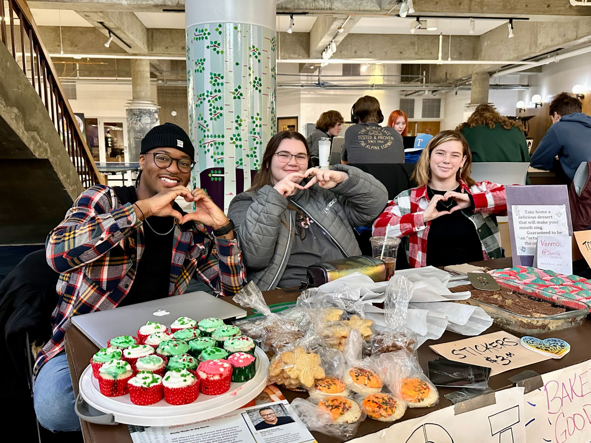 Students in the UAF Wood Center at a bake sale booth makes hearts with their hands