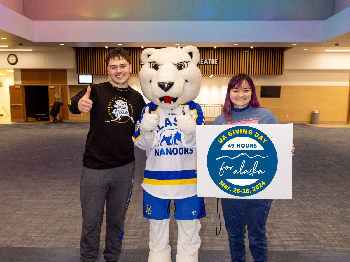 Students pose with Nook mascot in UAF Regents Great Hall holding sign with Giving Day logo