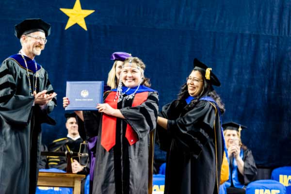 A Ph.D. student displays their diploma cover after recieving their hood from the UAF chancellor and provost.