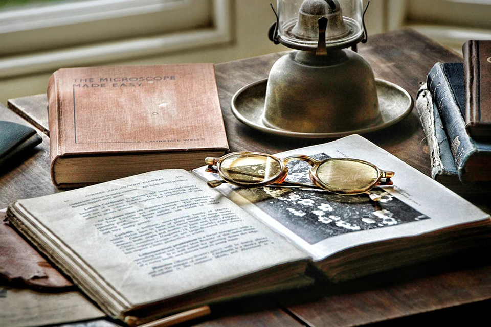 A book sits open with a pair of vintage glasses folded on top. An oil lamp sits on the desk behind them. | Stock image from Canva