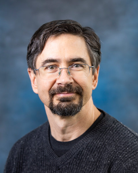 A portrait of a man with dark hair and glasses wearing a dark gray sweater