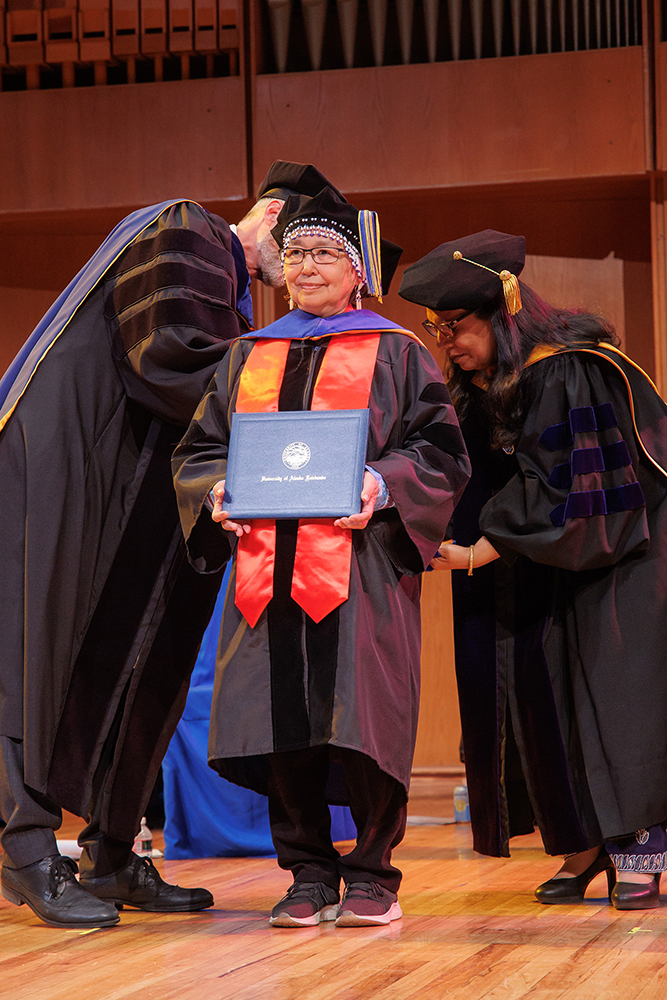Catherine Mary Moses stands while UAF Chancellor Dan White, left, and Provost Anupma Prakash perform the formal hooding during ceremonies in the Davis Concert Hall on May 5. Moses earned her Ph.D. in Linguistics: Interdisciplinary Studies. | Photo by Todd Paris