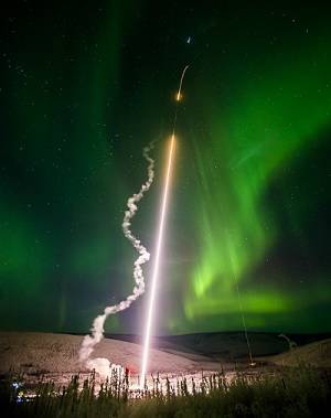 A rocket being launched into the night sky. Green aurora lights are visible in the background, and a trail of smoke shows the path of the rocket.
