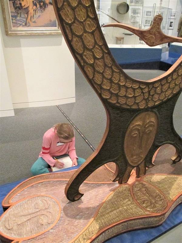 A child sits in a museum gallery in front of a wooden sculpture, sketching on a pad of paper in their lap.