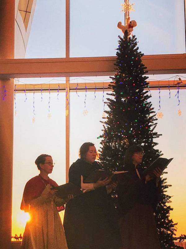 Three people dressed in 19th century style clothing, standing in front of a holiday tree in the museum lobby, holding songbooks and singing together.