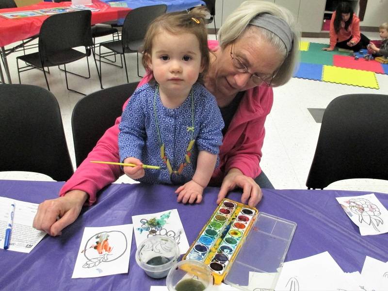 A child sits on an adult's lap, holding a paintbrush. A set of watercolor paints is on the table in front of them.