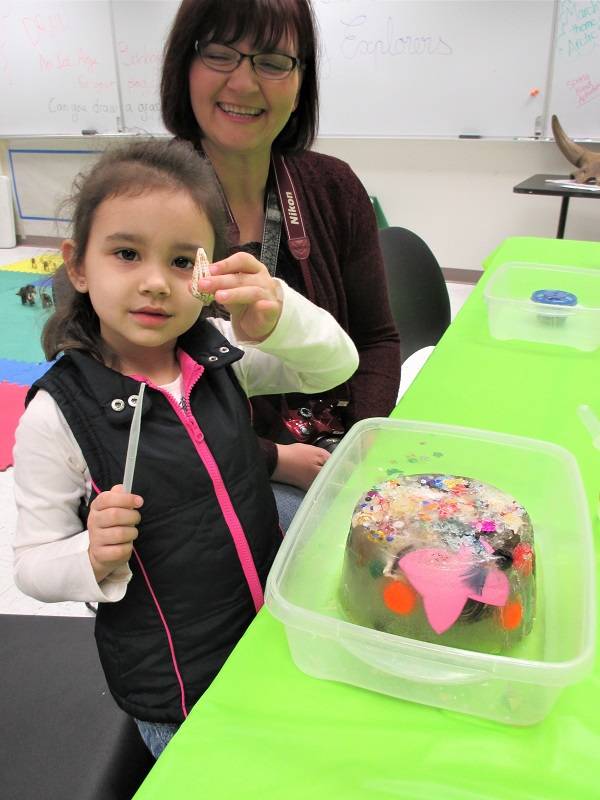 A child holding a shell and a pipette stands next to a block of ice with sequins and other shiny items frozen inside.