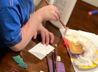 A child holds a colored pencil. A variety of craft materials are laid out on a table in front of the child.