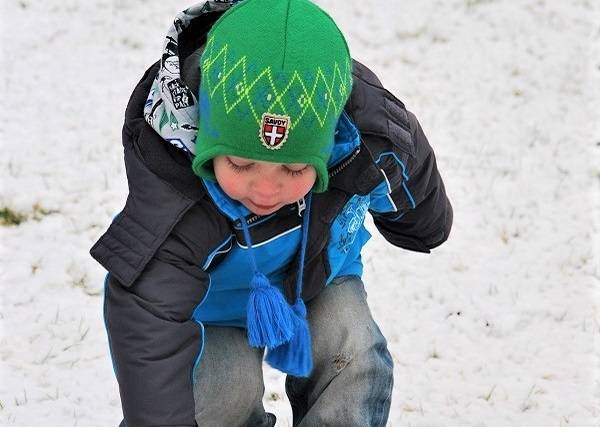 Child wearing a blue jacket and green hat bending down to play in the snow.