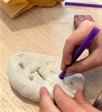 Child's hands holding a purple straw, making designs on a piece of white clay.