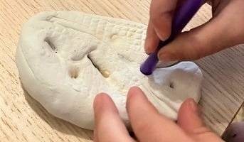 Child's hands holding a purple straw, making designs on a piece of white clay.