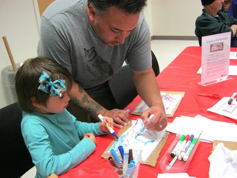 An adult stands behind a child sitting at a table. Both are using markers to color a model of a watershed.