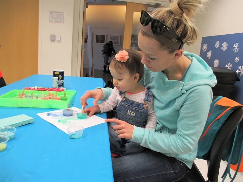 An adult sits with a child on their lap, helping the child paint with colored ice cubes.