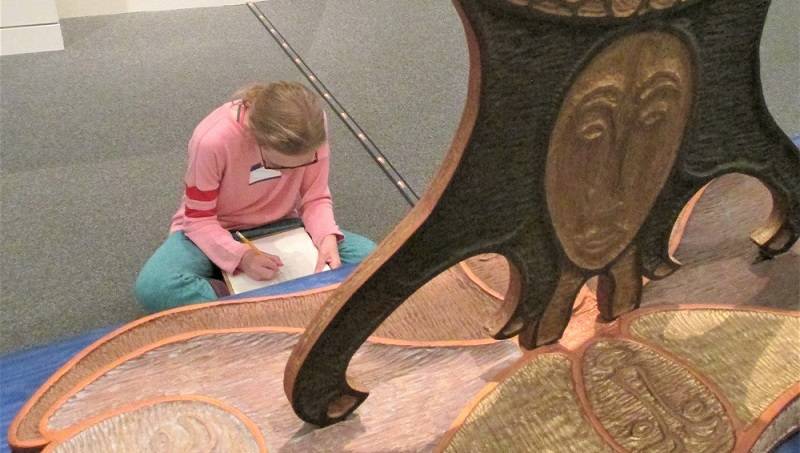 Child sitting in a museum gallery, sketching on white paper. A wooden Raven sculpture is in front of the child.