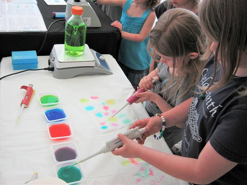 Two children stand at a table pipetting colorful liquids onto paper towels.