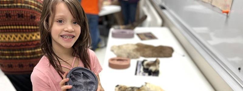 Child holding a mold of a wolf track, standing next to a table with a wolf pelt and other museum objects.