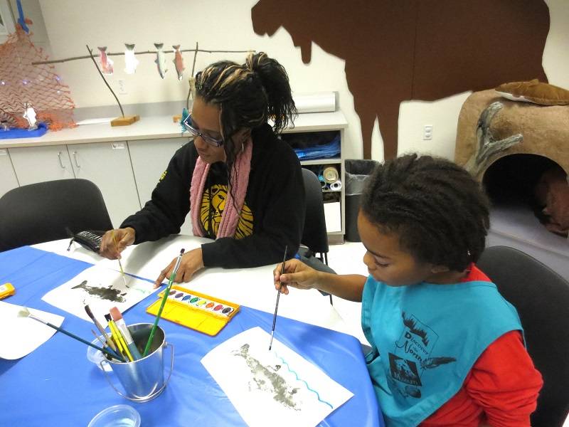 An adult and a child sit at a table, painting pictures of fish with watercolors.