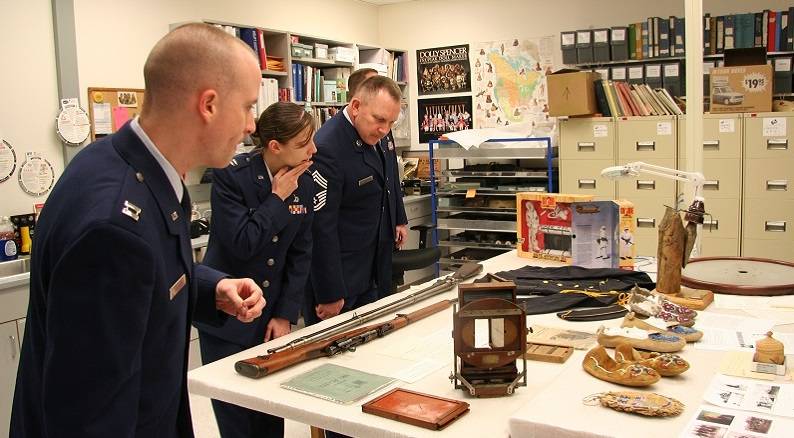 Three people wearing military uniforms looking at a variety of museum objects on a table.