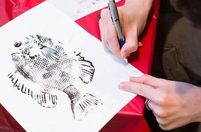 A hand holding a marker and drawing details on a fish print. A replica fish covered in ink is visible in the background.